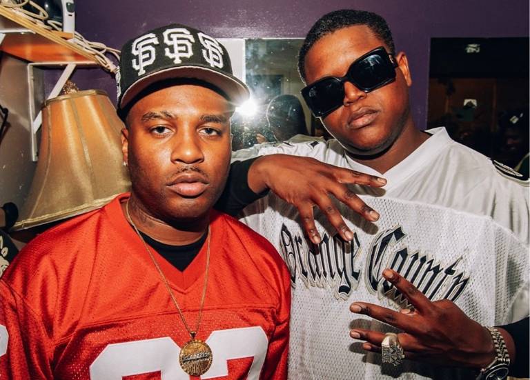 Two Black men in sports jerseys look into the camera as a flash is reflected in a mirror behind them