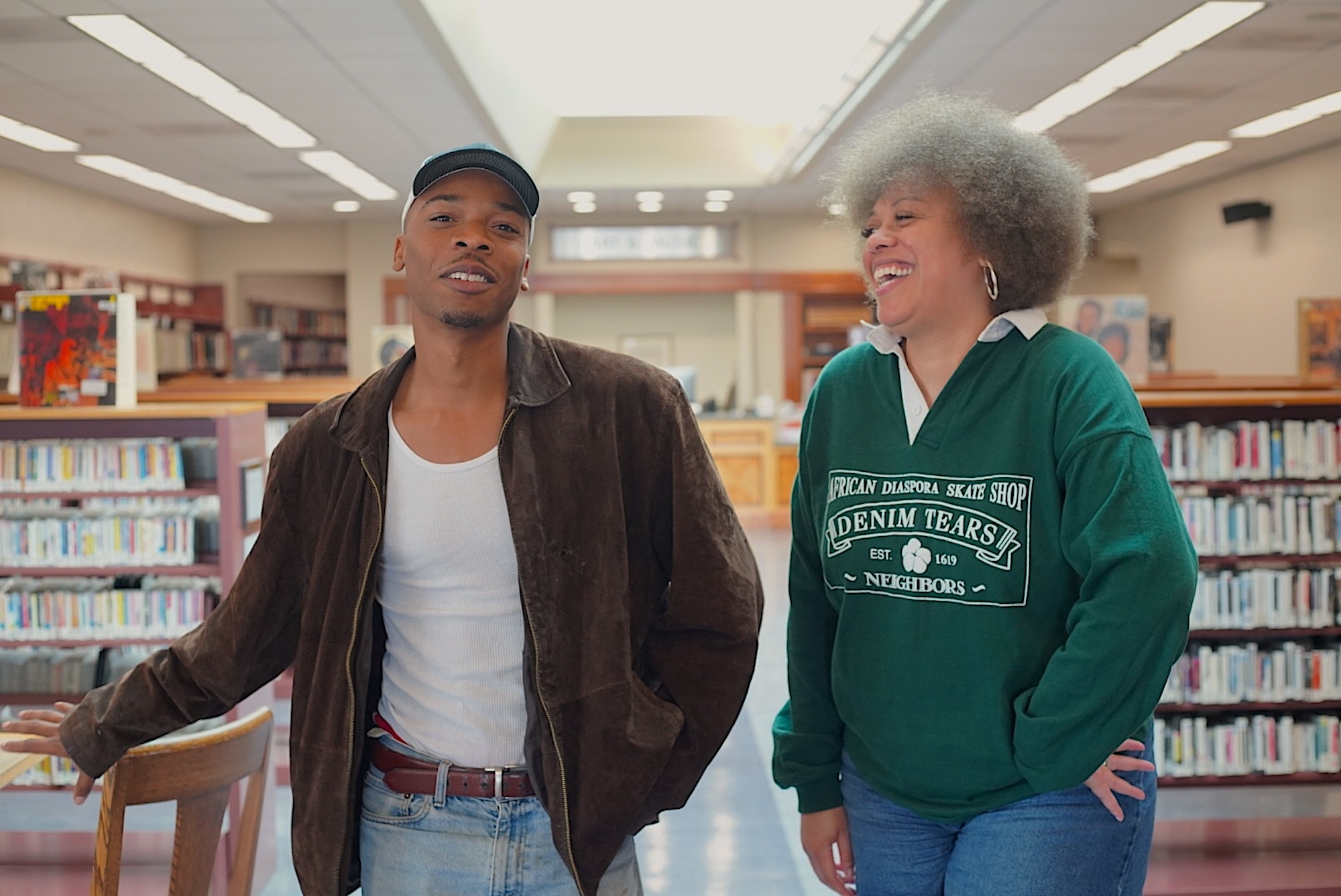 An African American man in a hat and brown jacket and a woman in a green shirt and large afro pose for a photo inside of a library.