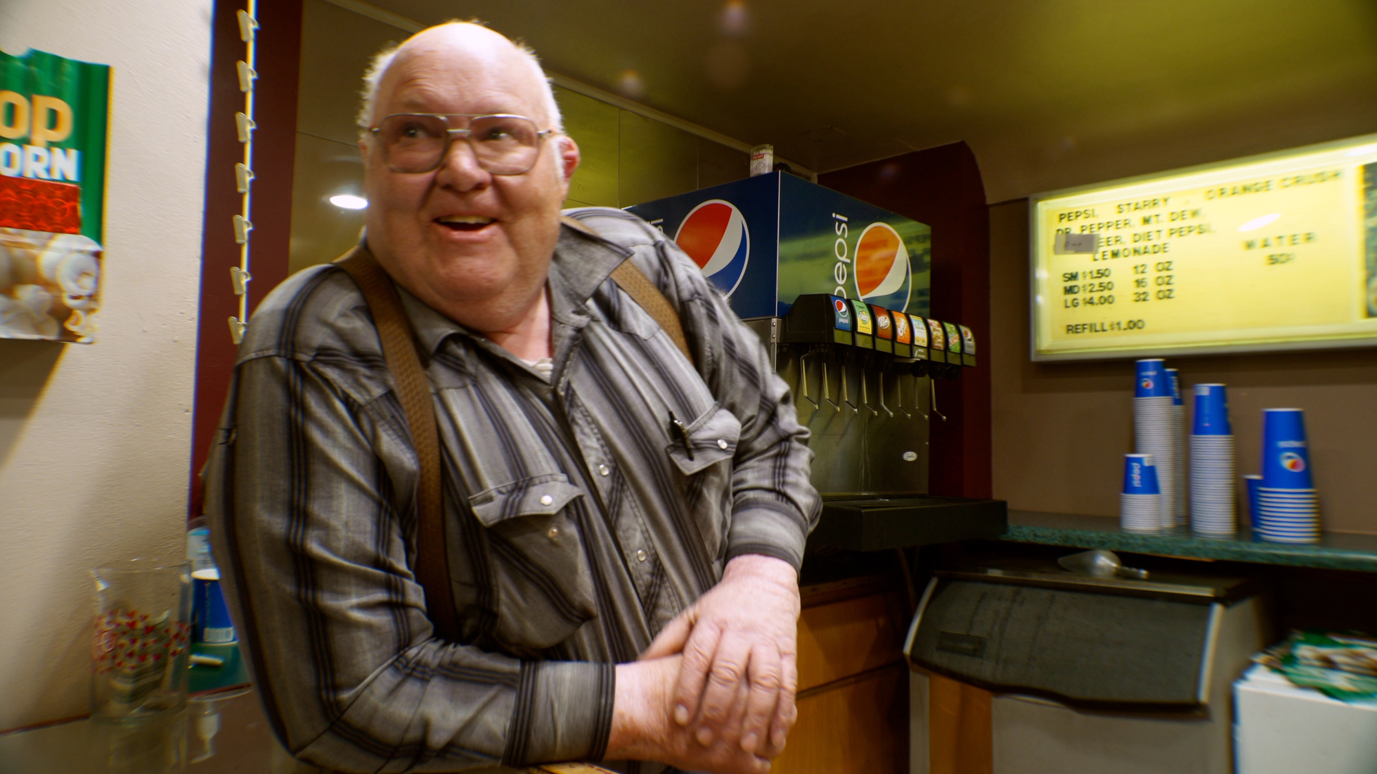 older white man in suspenders leans against concession stand counter