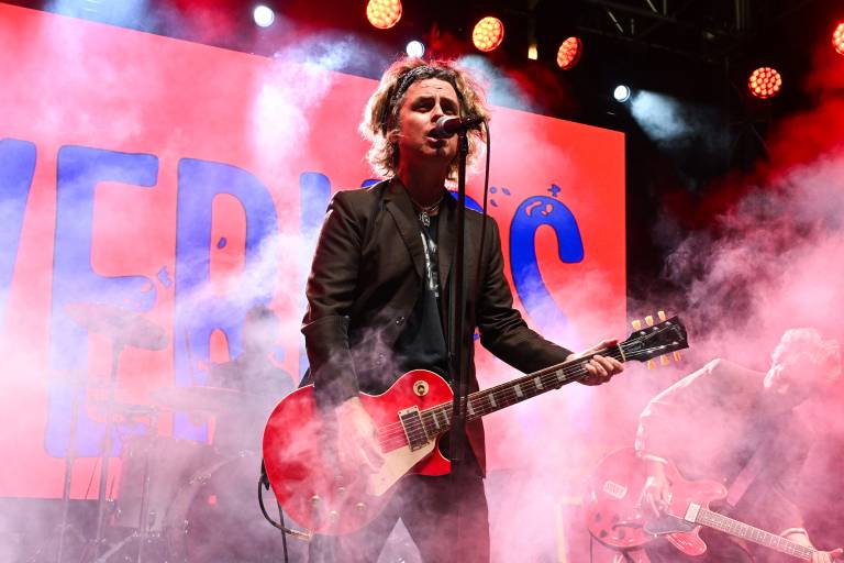 A man in a black suit jacket an shabby hair plays a red guitar against a backdrop of smoke and a red screen