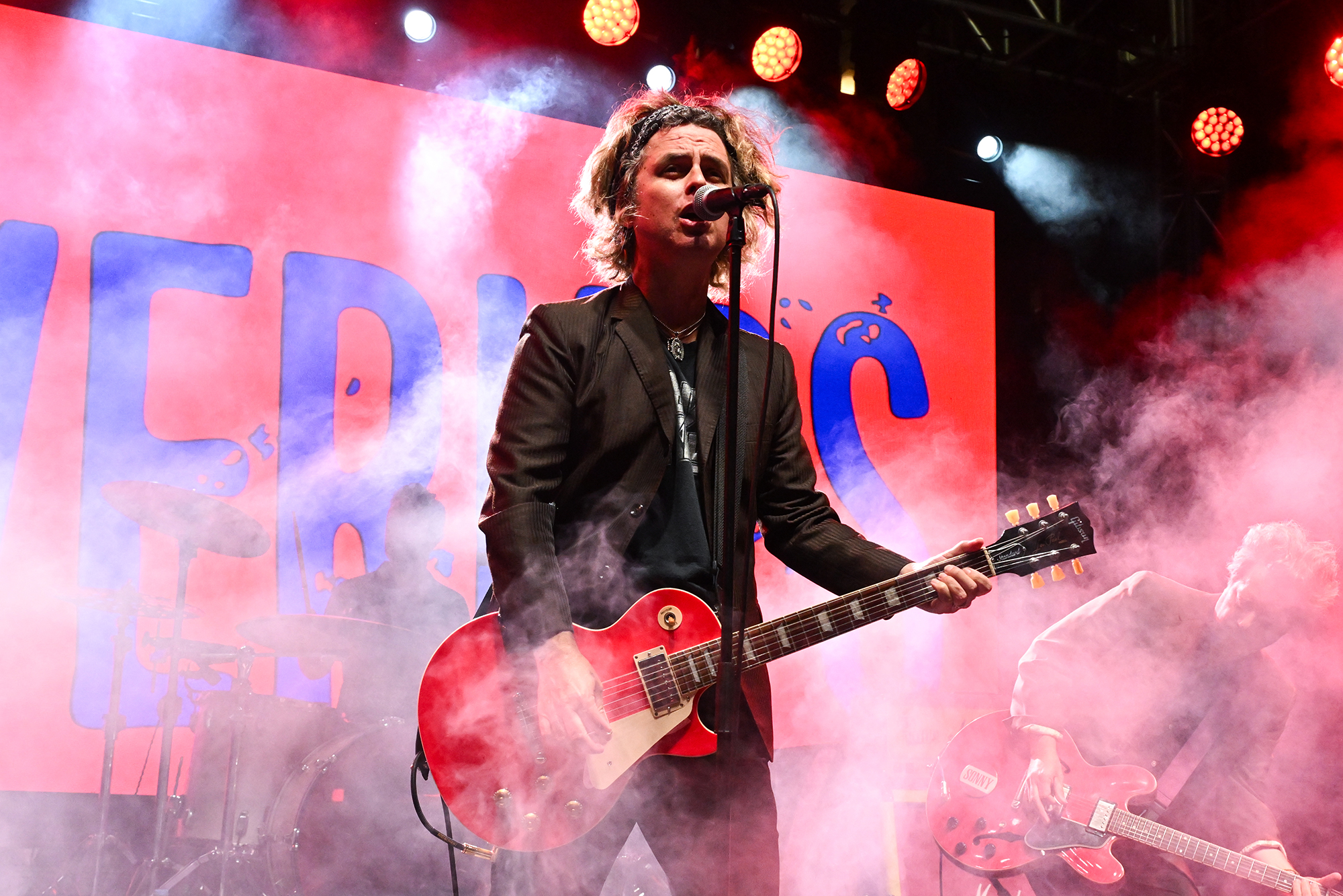 A man in a black suit jacket an shabby hair plays a red guitar against a backdrop of smoke and a red screen