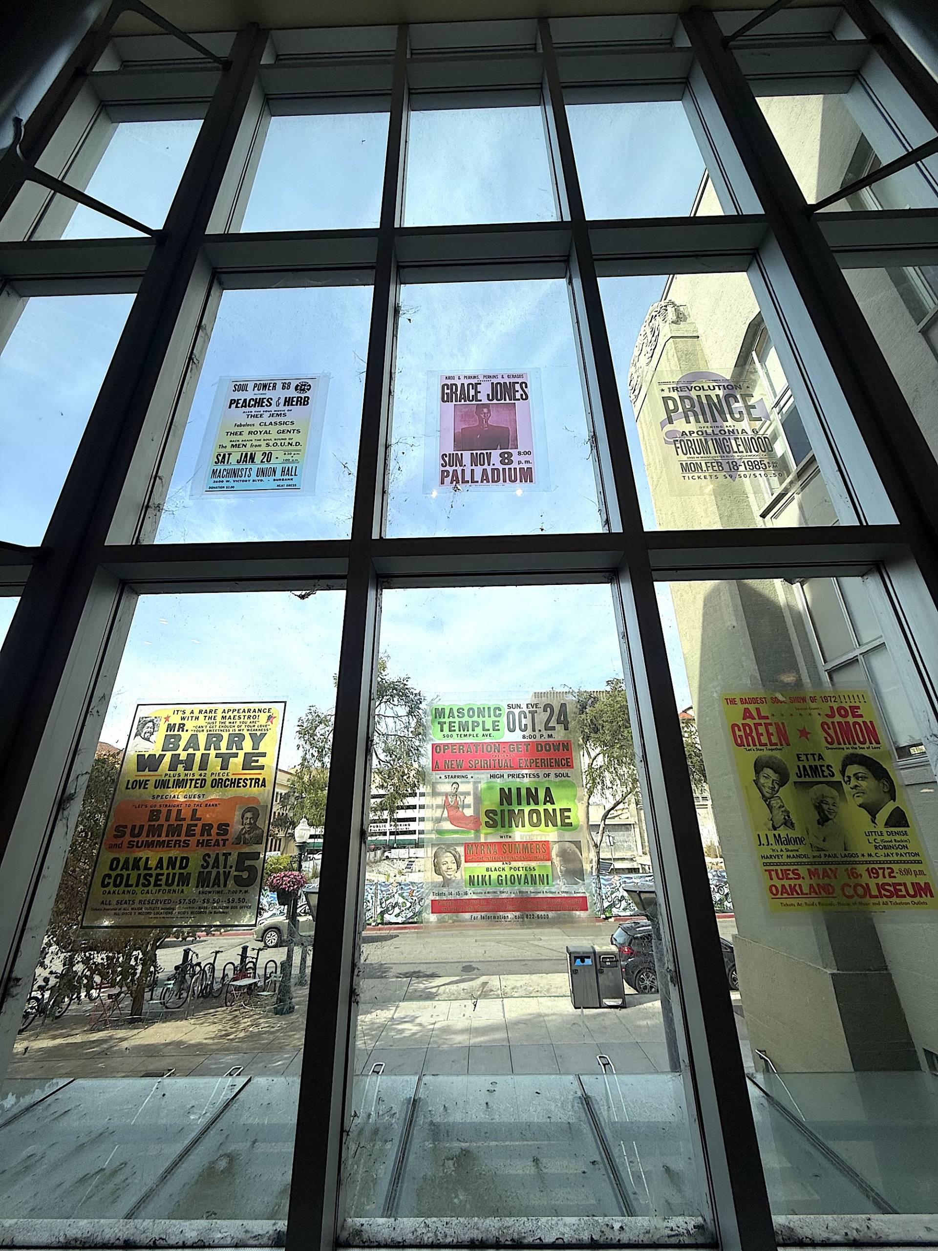 Looking out the window from inside of the Main branch of the Berkeley Public Library, as translucent concert posters adorn the windows.