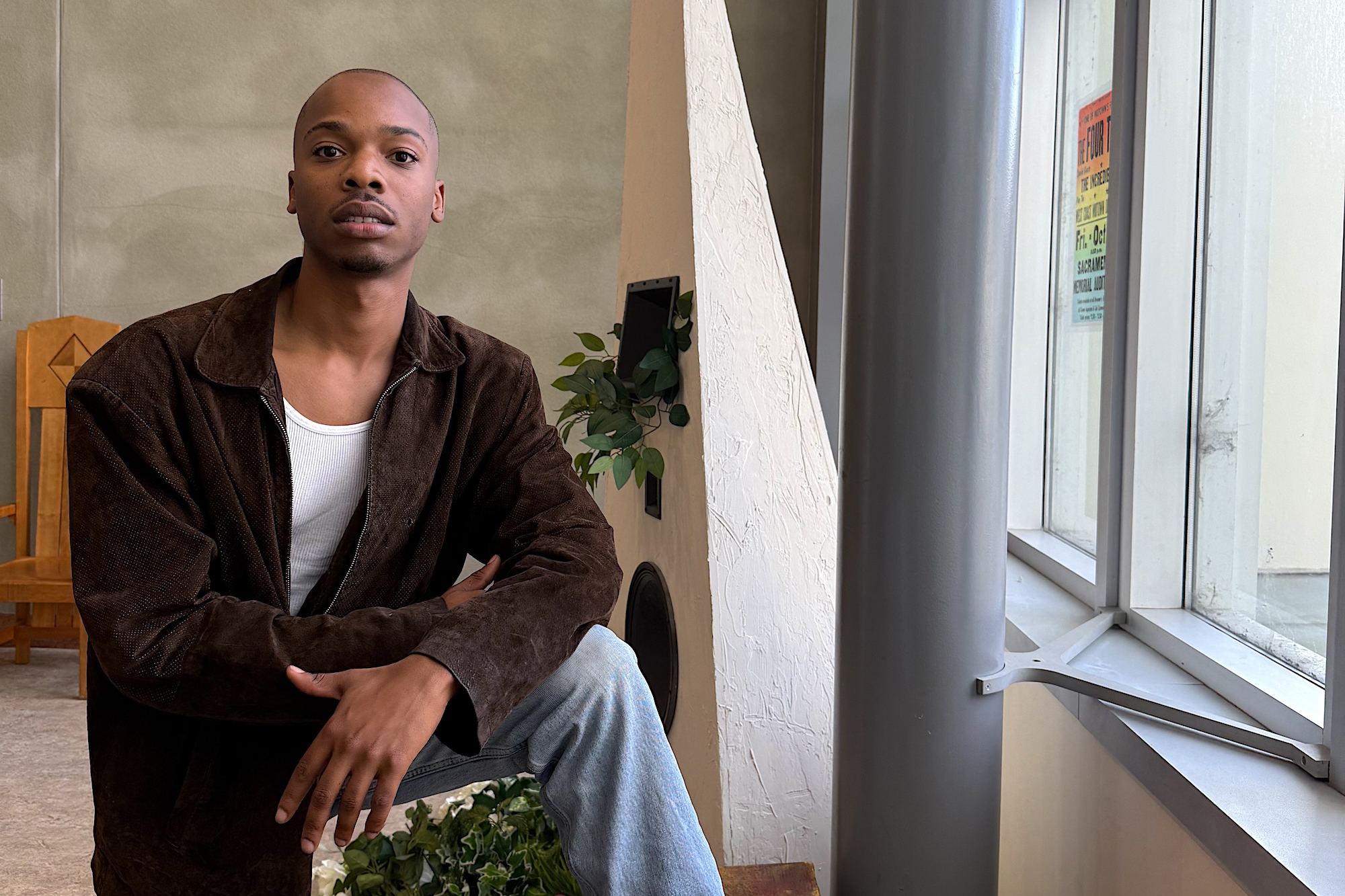 A young Black man poses in a brown jacket and jeans near a sunlit window