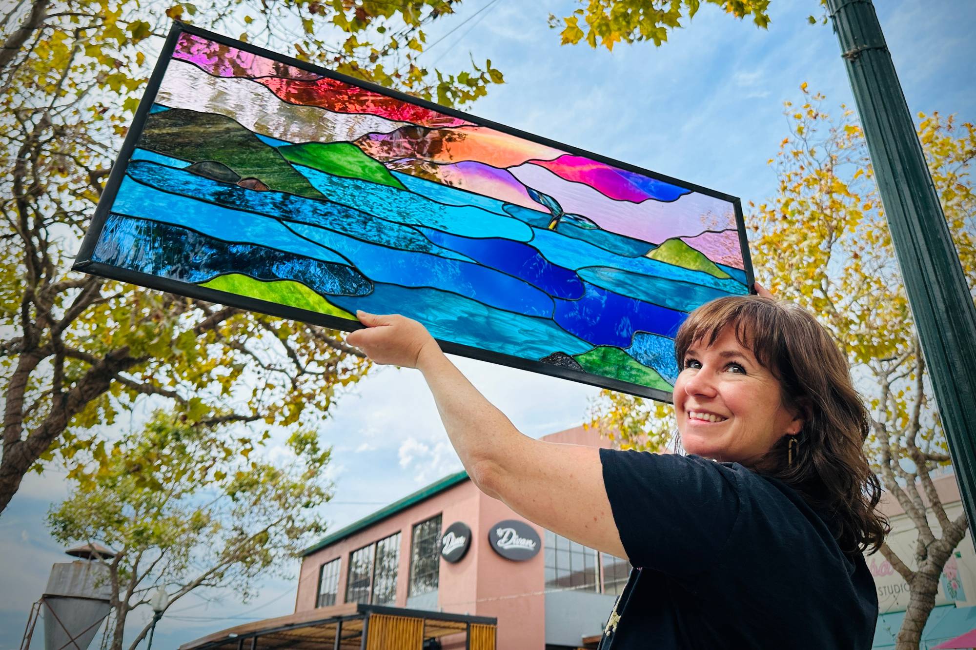 A woman in a black top turns and smiles at the camera while holding up a stained glass window showing the form of a sunset over the ocean