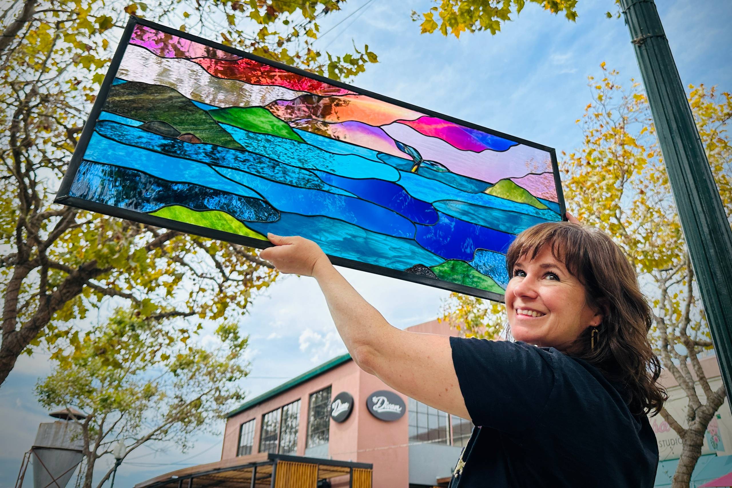 A woman in a black top turns and smiles at the camera while holding up a stained glass window showing the form of a sunset over the ocean