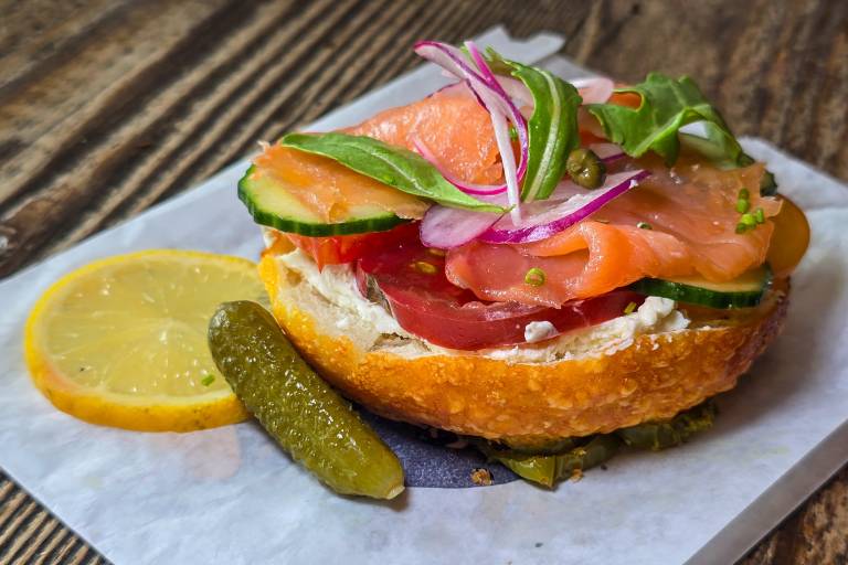 A halved bagel topped with cream cheese, tomato, red onions and lox with some cornichons on the side.