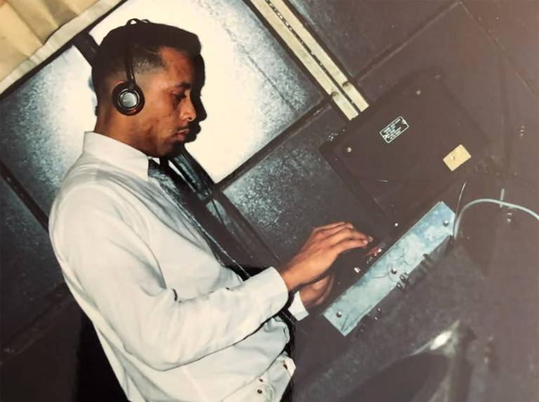 A young Black man in a white shirt and tie wears headphones as he focuses on a turntable and mixer