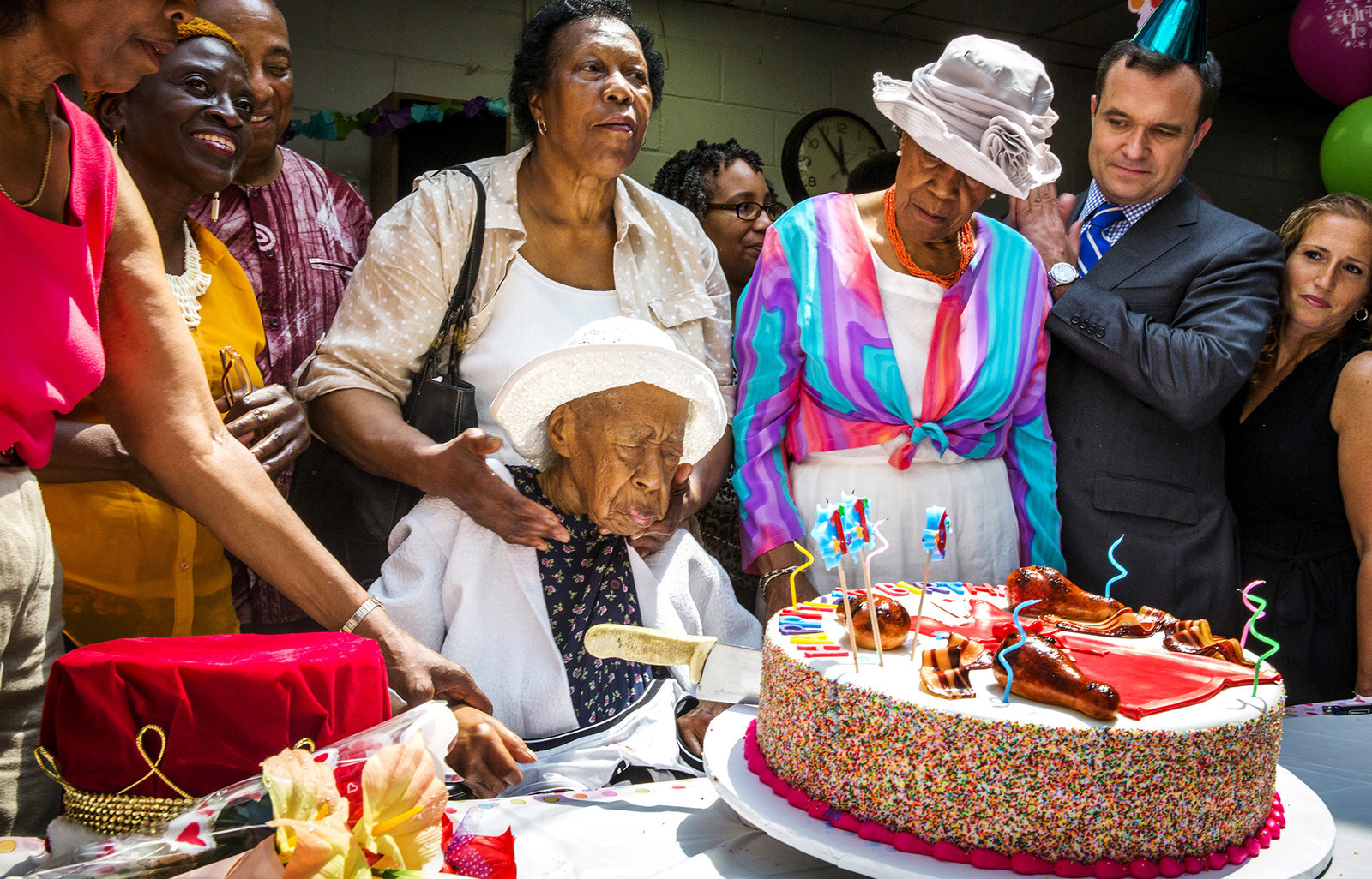 old Black woman faces large birthday cake with poeple around her