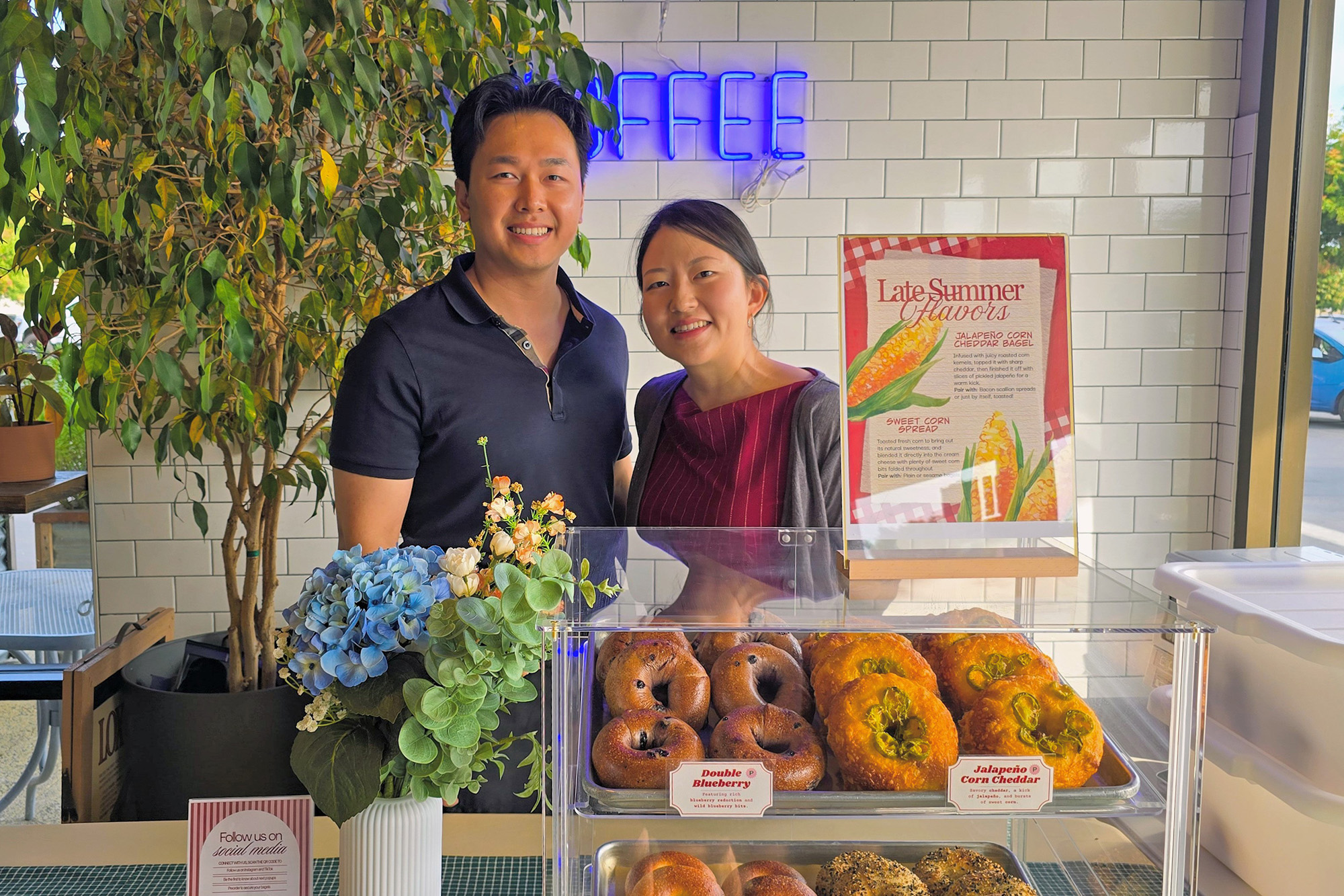Asian man and woman pose for a portrait in front of a display of bagels.