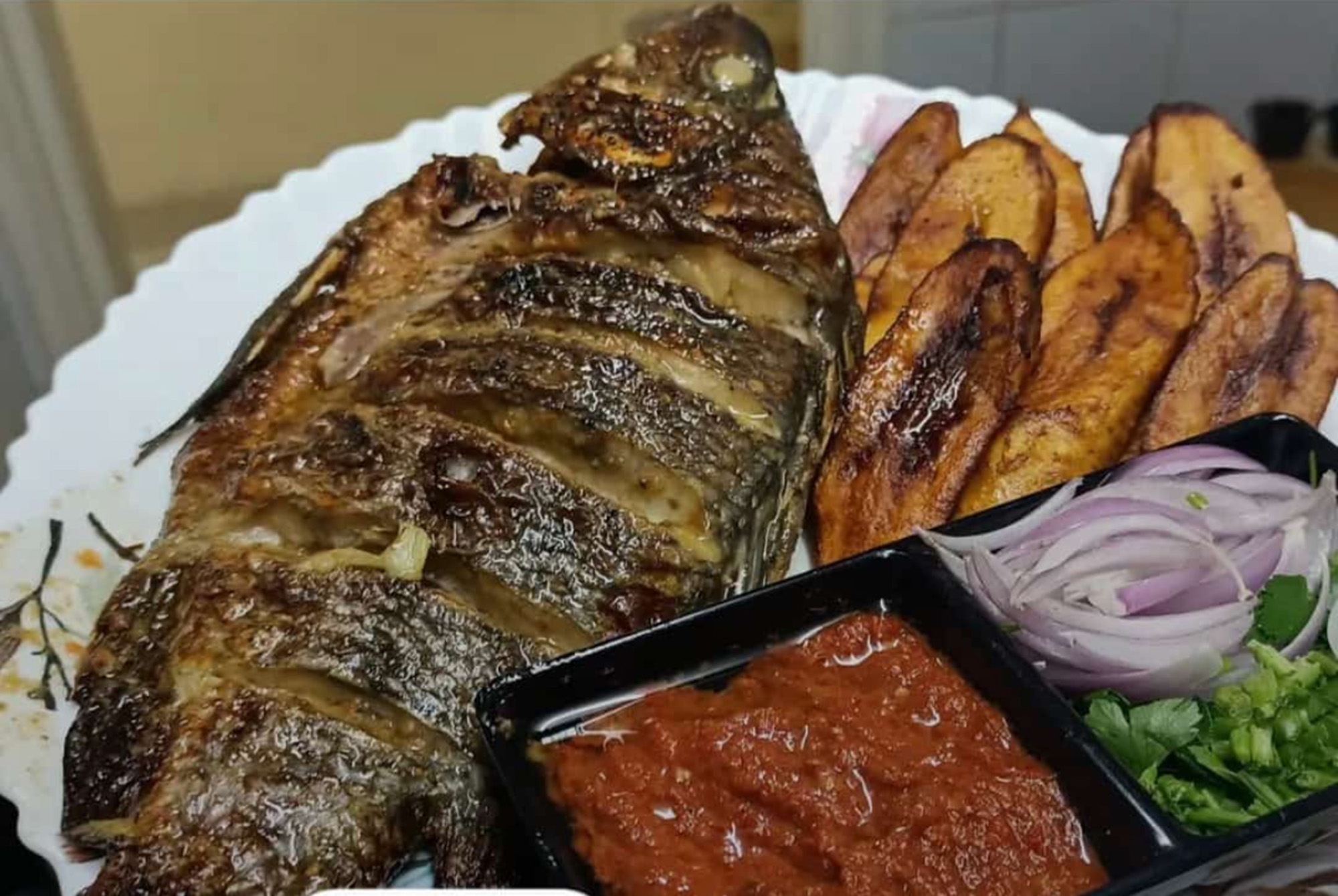 Grilled fish, fried plantains, hot sauce on a plate.