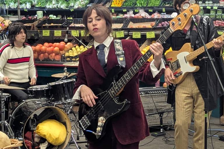 A rock band performs in the produce section of a grocery store. In front, the bass player wears a red velvet suit.