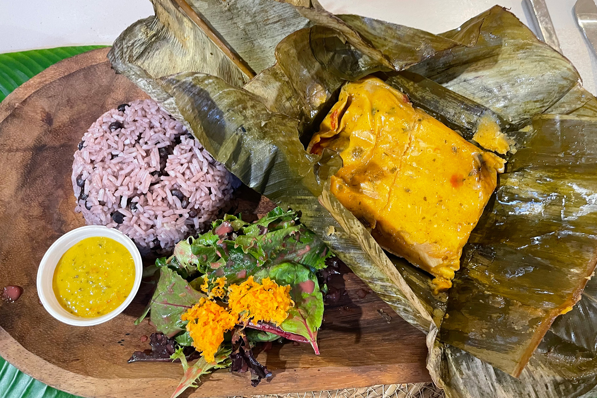 A wooden platter with a tamal, rice and beans, and a mustard-yellow salsa.