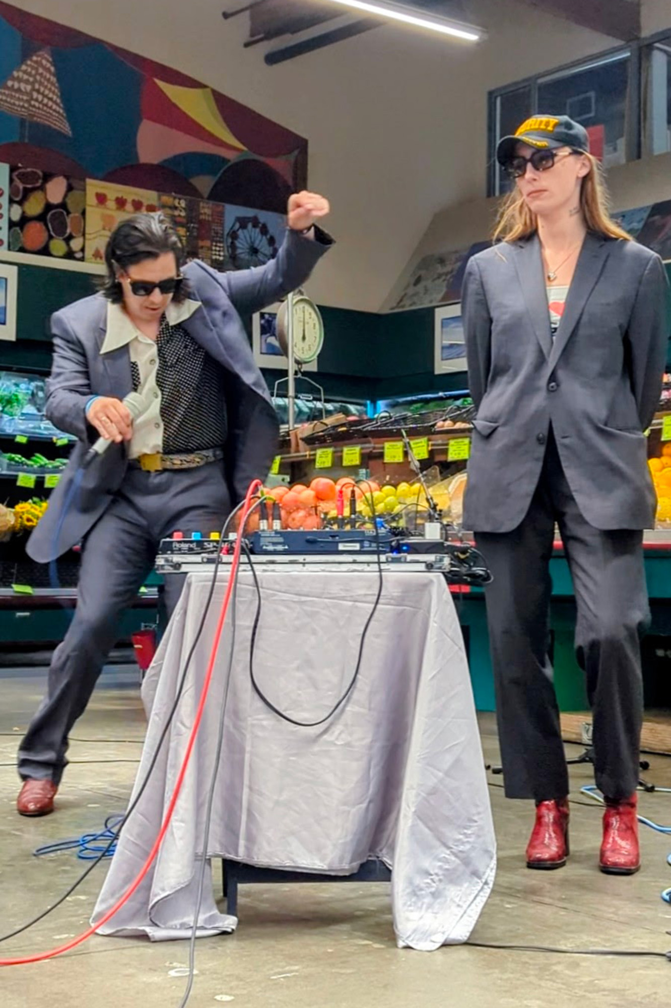 Two musicians in gray suits performing in the produce section of a grocery store. The man in sunglasses on the left dances energetically. 