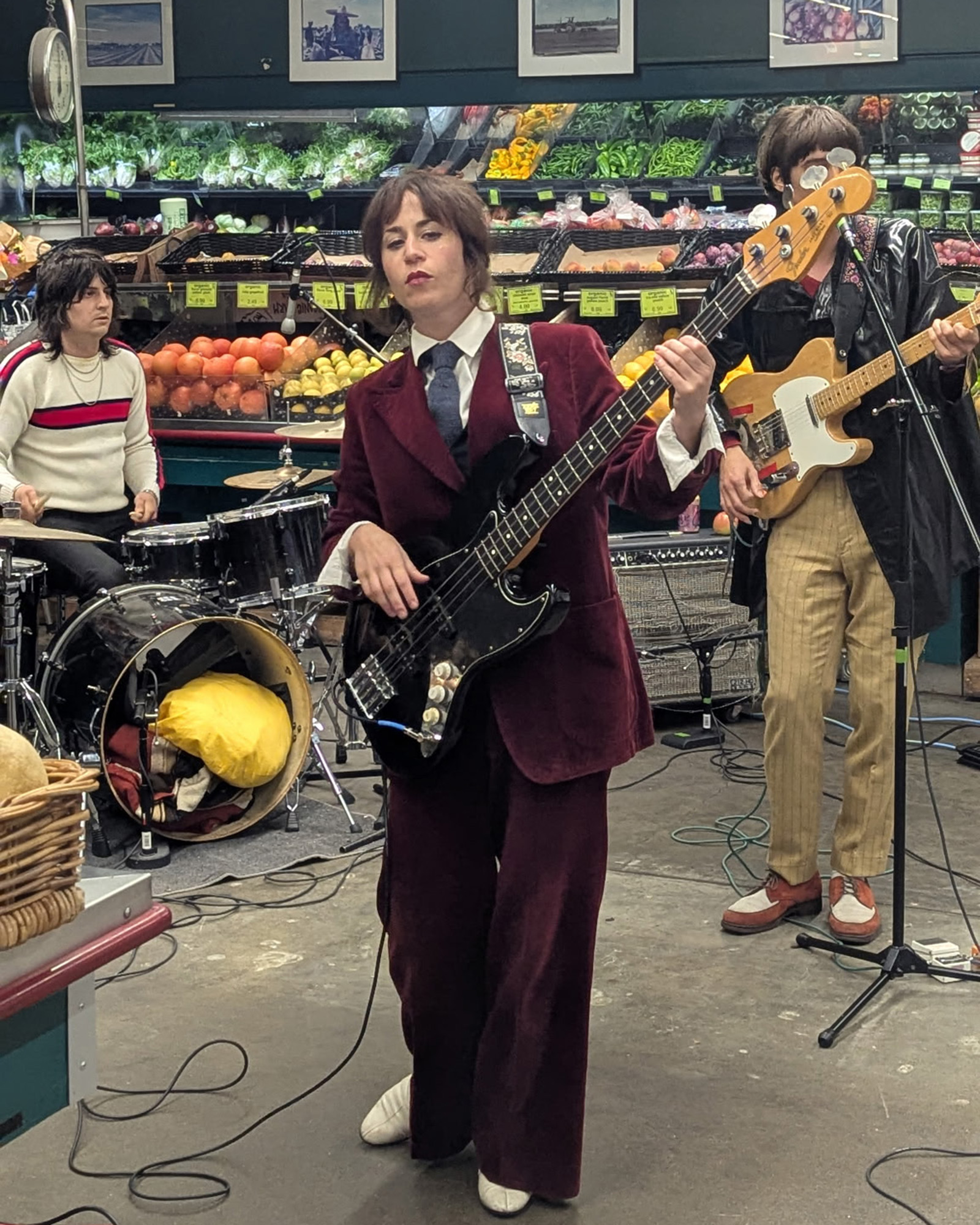 A rock band performs in the produce section of a grocery store. In front, the bass player wears a red velvet suit.