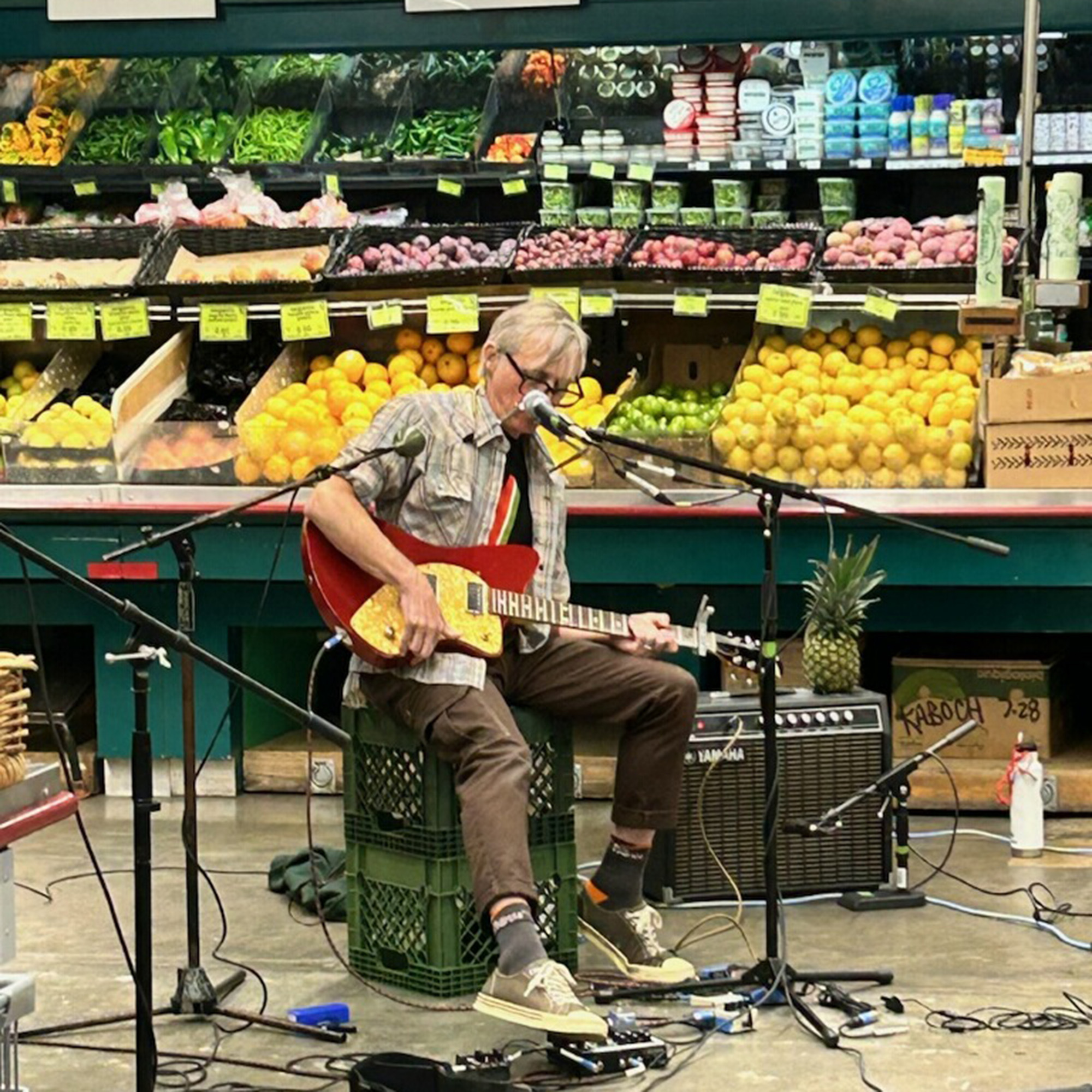A musician, seated in the produce section of a grocery store, plays the guitar.