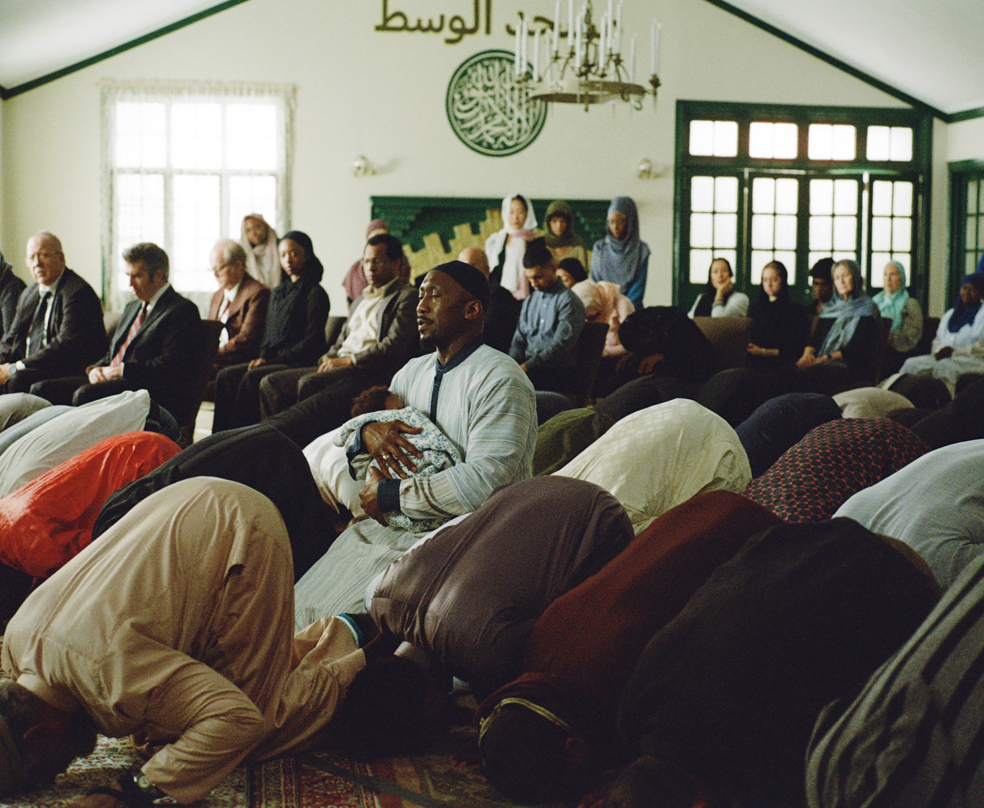 A Muslim man in a masjid holds a baby as others around him bow down in prayer.