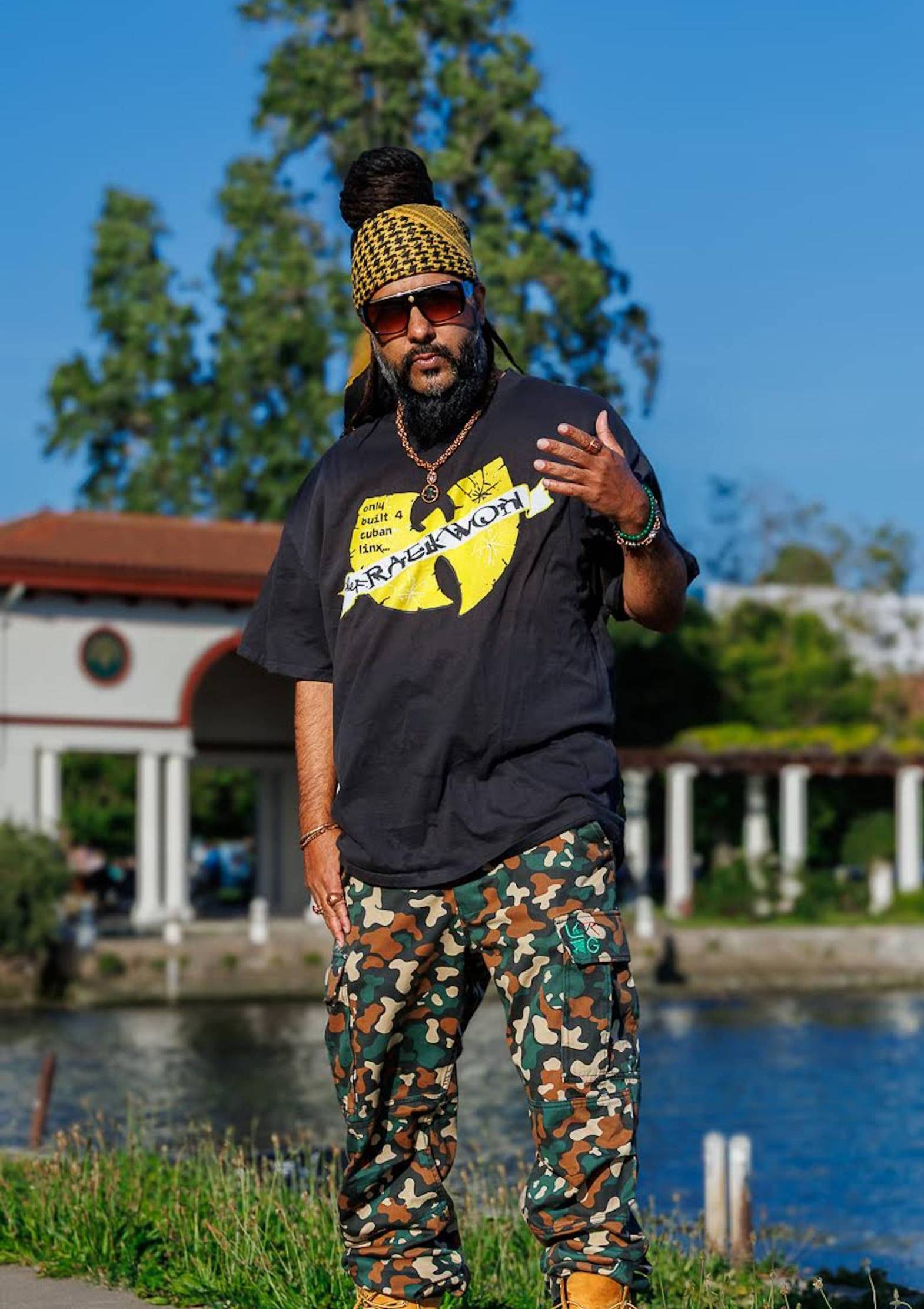 A man in sunglasses, black shirt and multicolored pants posing for a photo near a lake.