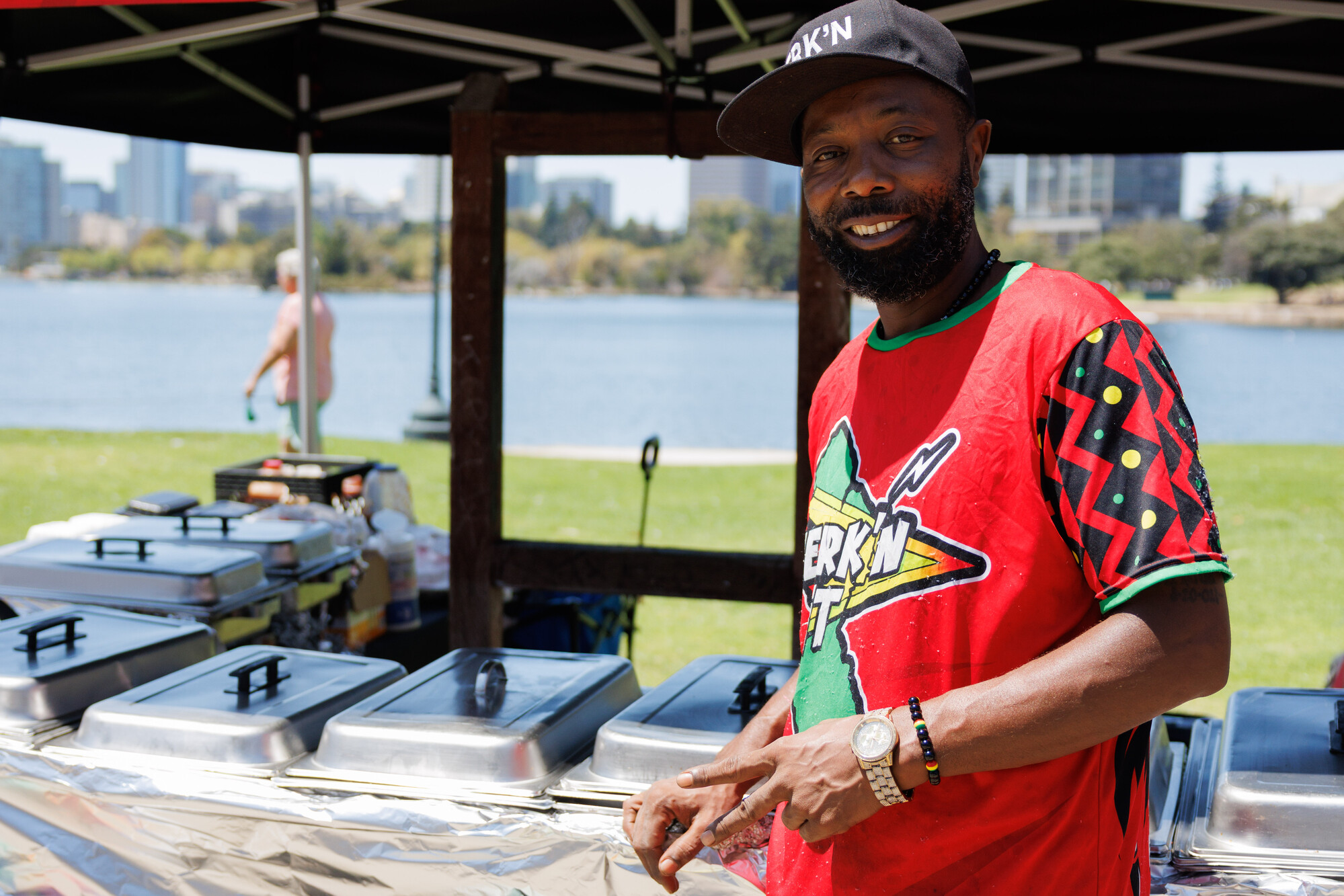 Man in a bright red T-shirt (the text reads, "Jerk'n It") poses for a portrait in front of a lakeside food stand.
