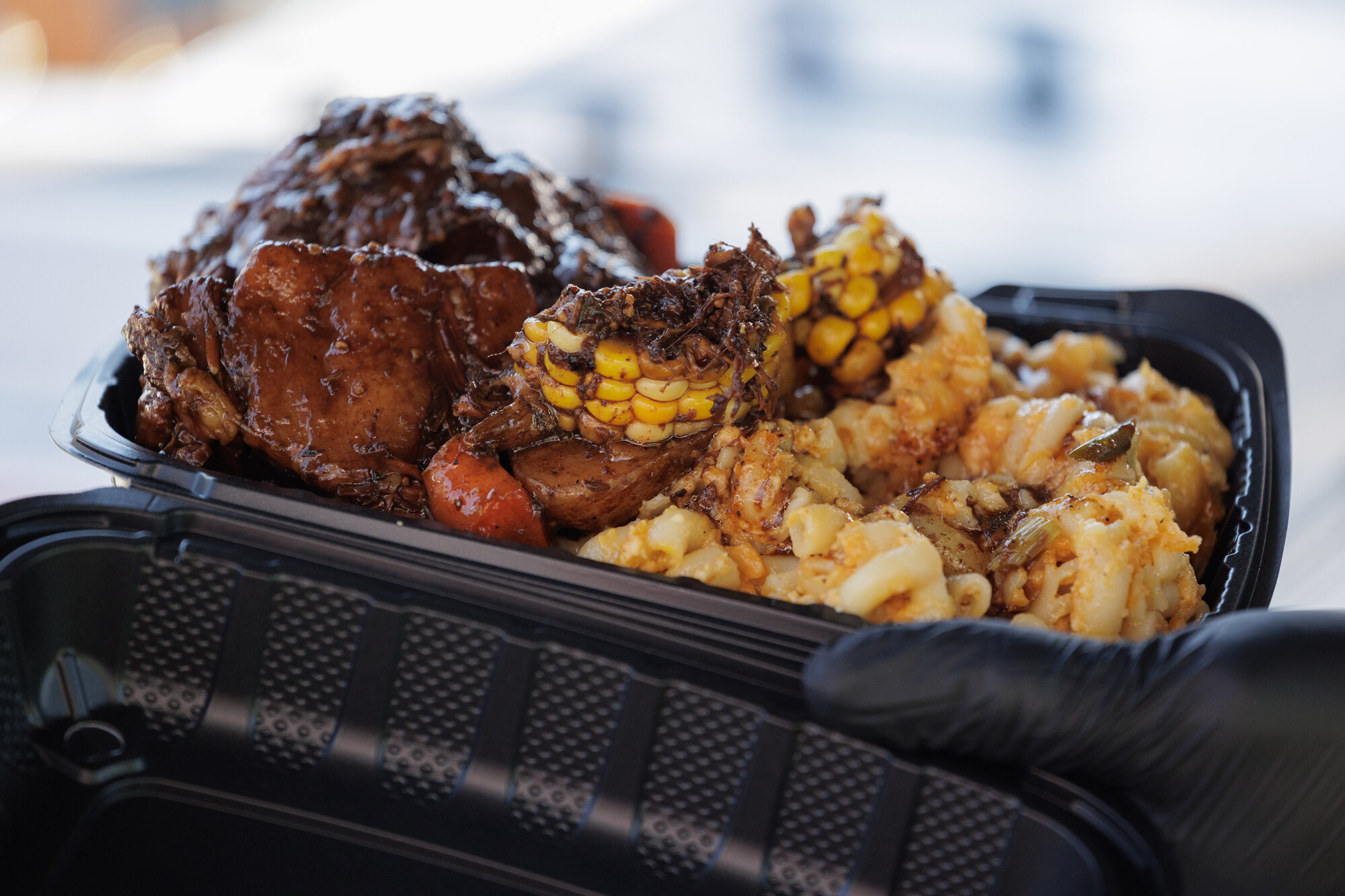Plastic takeout carton piled high with oxtails, corn on the cob, and mac and cheese.