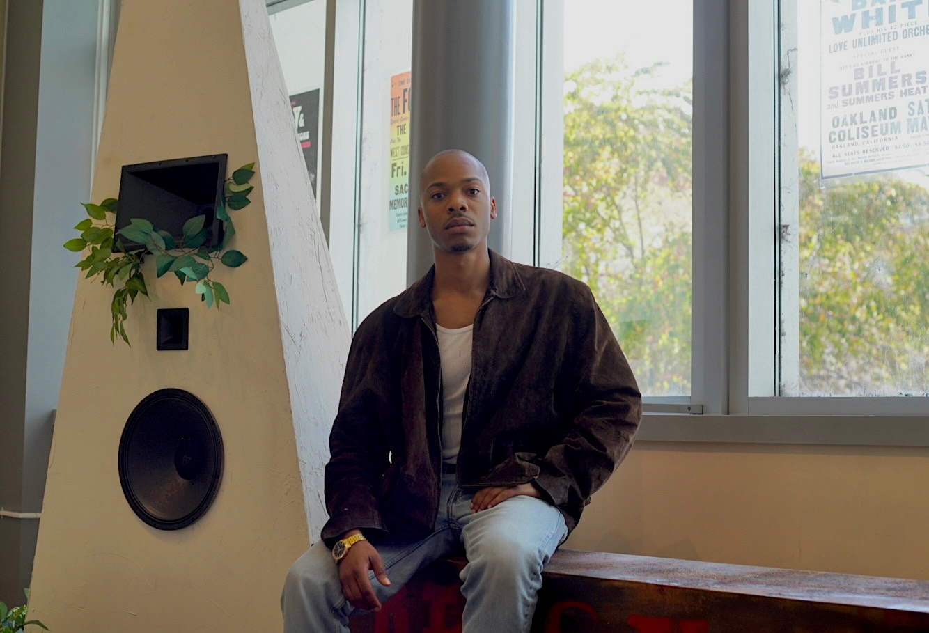 A young Black man poses in a brown jacket and jeans near a sunlit window