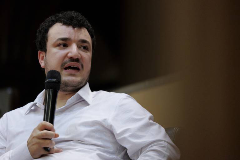 A man in a white collared button-down shirt holds a microphone and speaks, looking off to the right