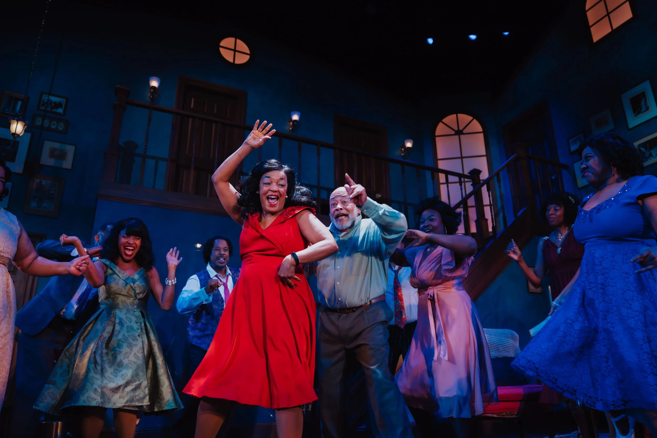A Black woman in a bright red dress smiles and dances in a dimly lit room with several others
