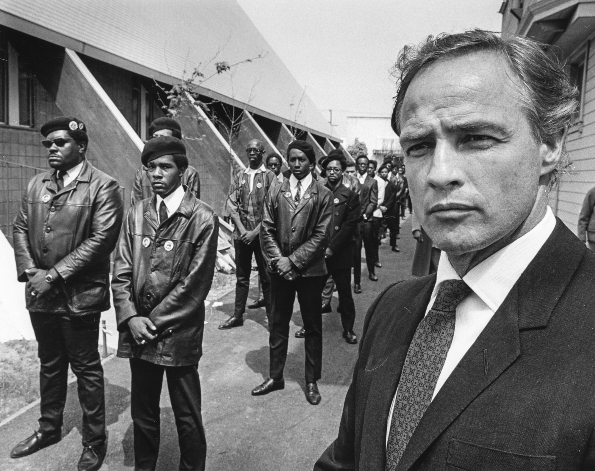 At right, a white middle-aged man in a suit and tie looks into the distance; at left, more than a dozen Black men in leather jackets and berets stand in two single-file lines next to an A-frame building