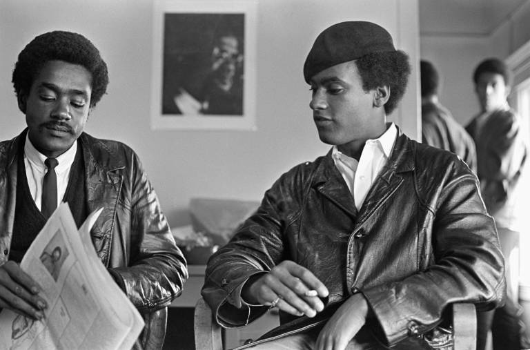 Two young Black men sit side-by-side in a room with white walls and a portrait of Malcolm X in the background