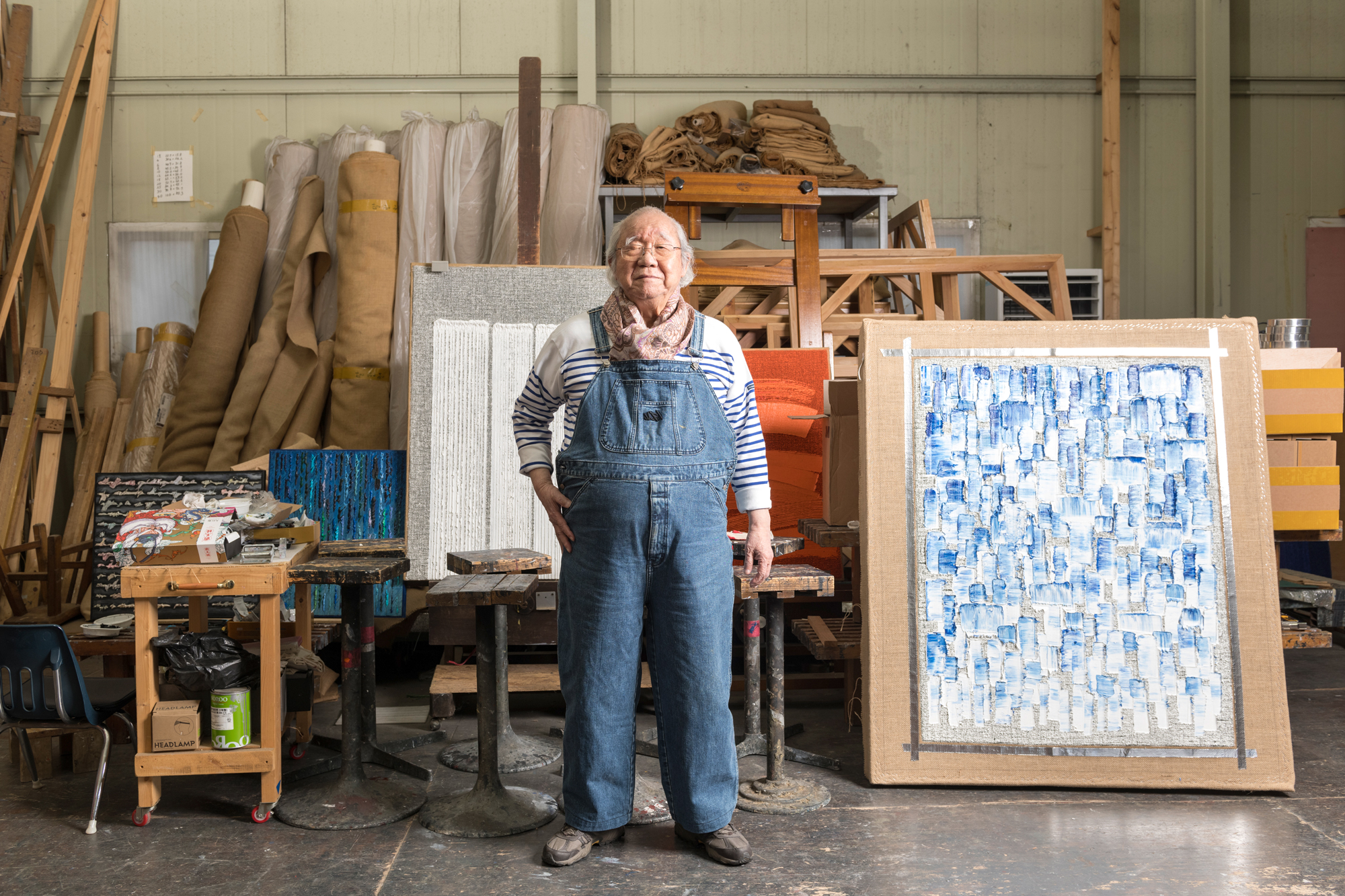 older Korean man in denim overalls next to abstract painting