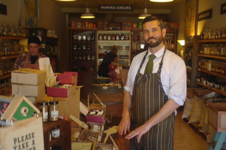 Bearded an in an old-timey apron poses for a portrait inside a well-stocked spice shop.