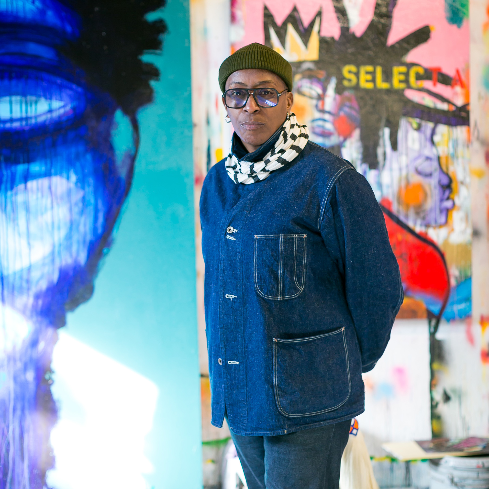 A woman stands in front of a Basquiat-style mural while wearing a blue chore coat, green beanie and black-rimmed glasses.