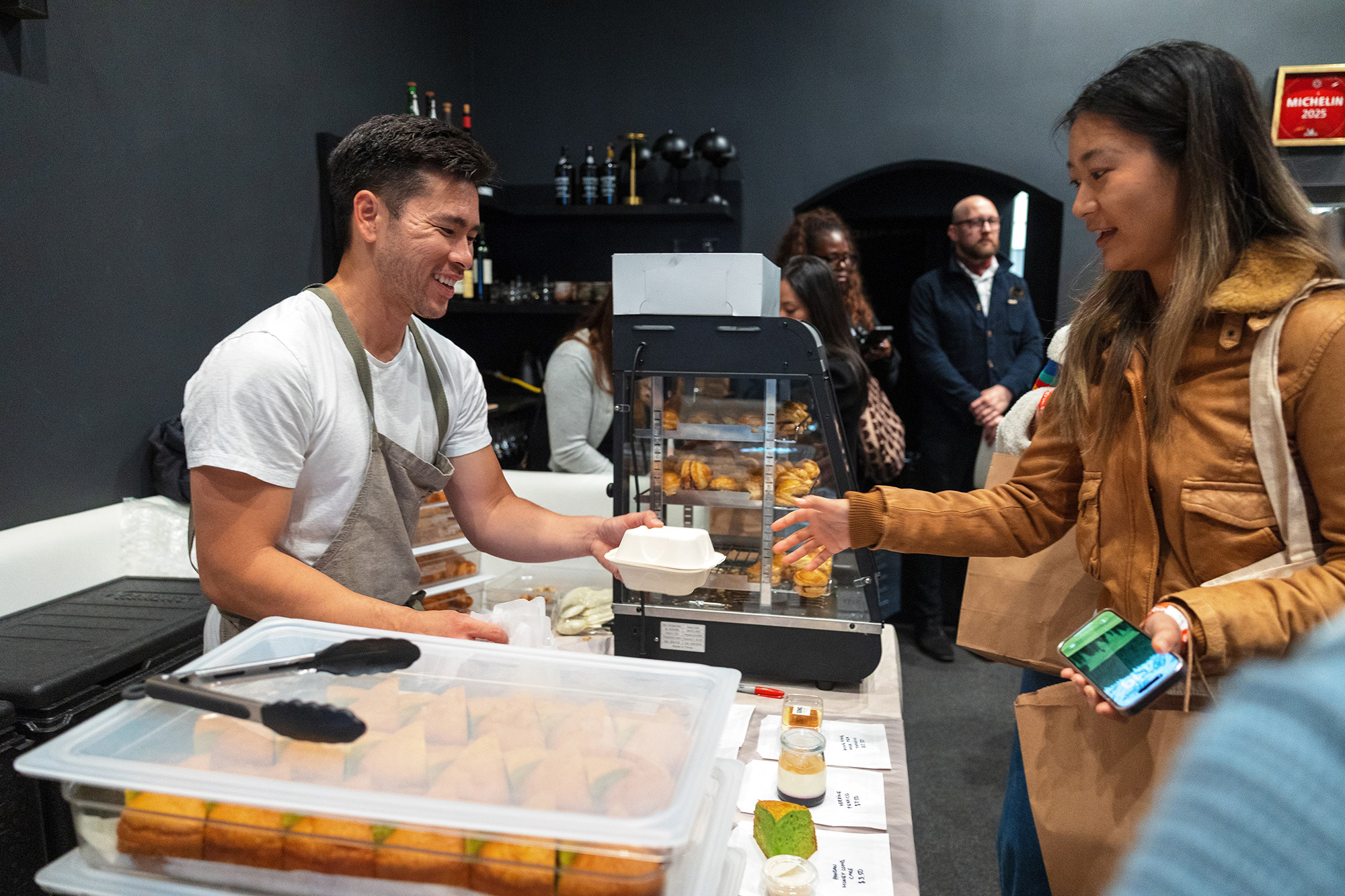 Asian man in a chef's apron hands baked goods to a customer.