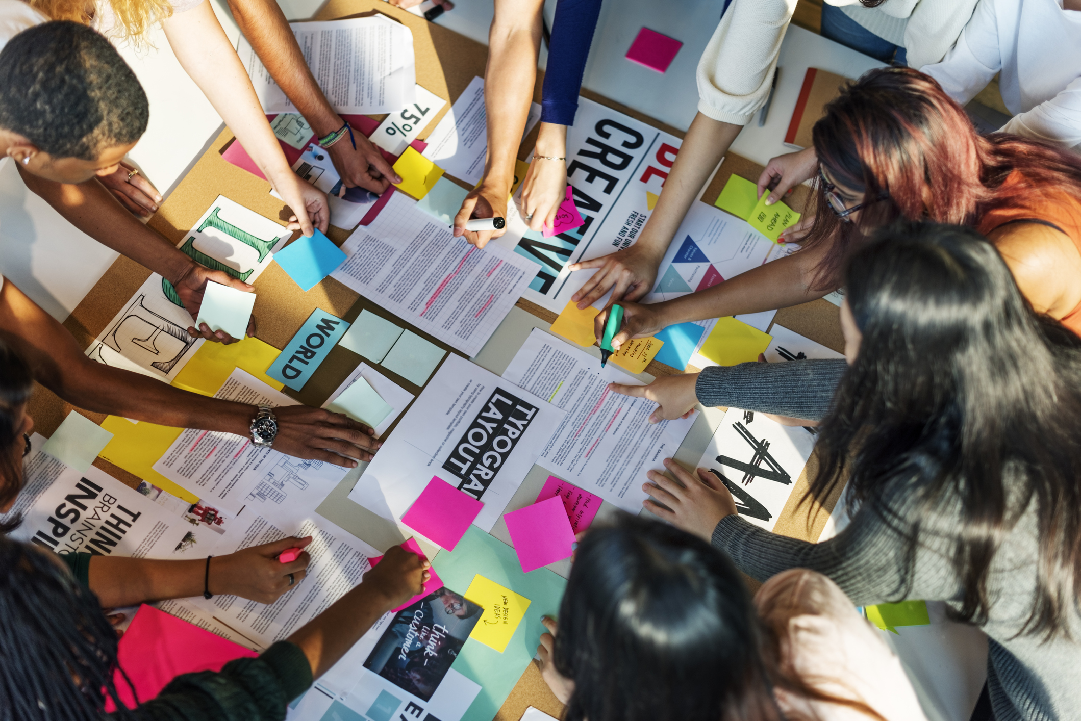 Above view of a group of students working around a large brown board covered in papers and labels. Hands of various skin tones reach into the center with highlighters, markers and sticky notes.