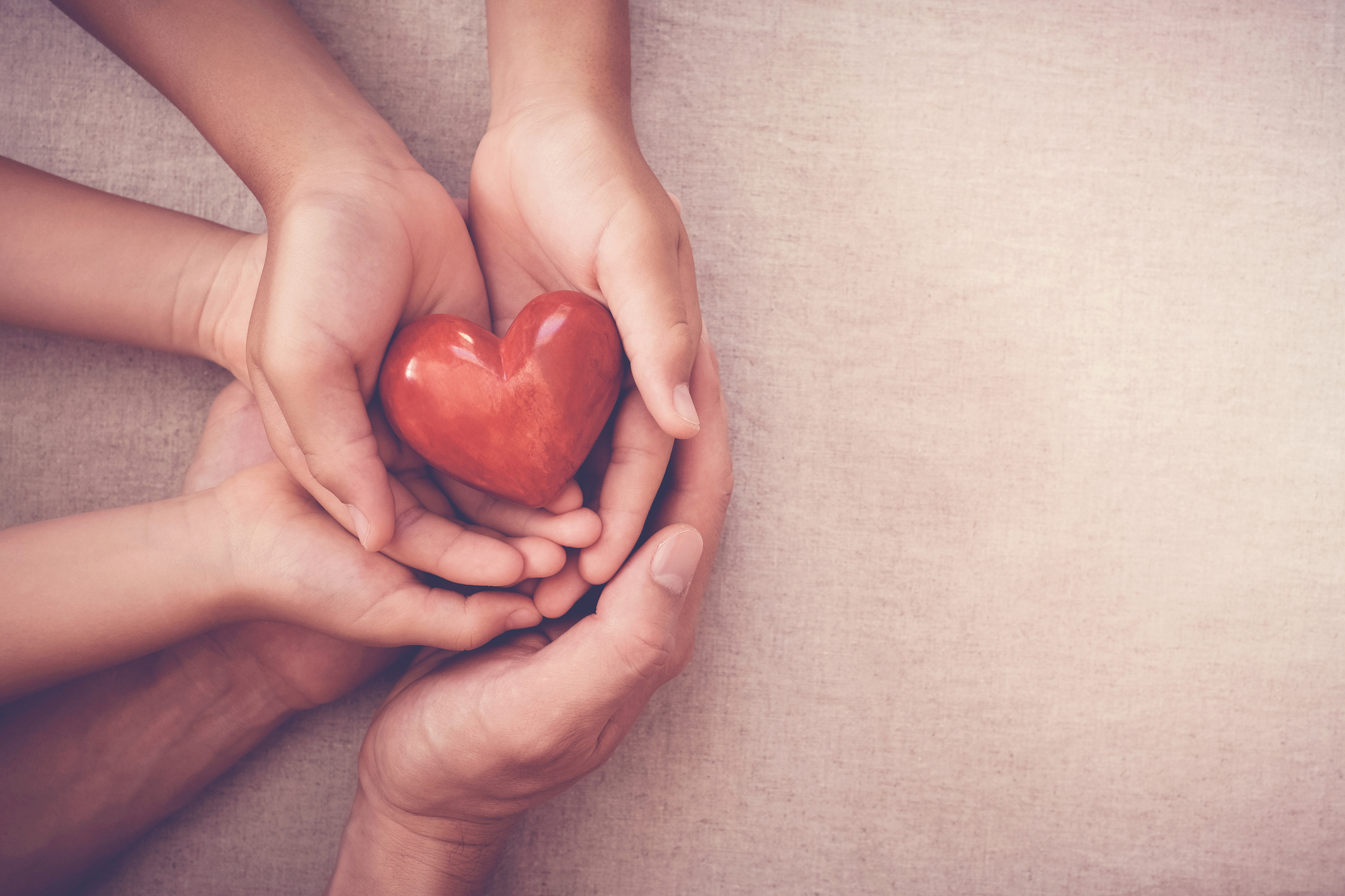 3 pairs of hands hold a red smooth stone heart. Beige background
