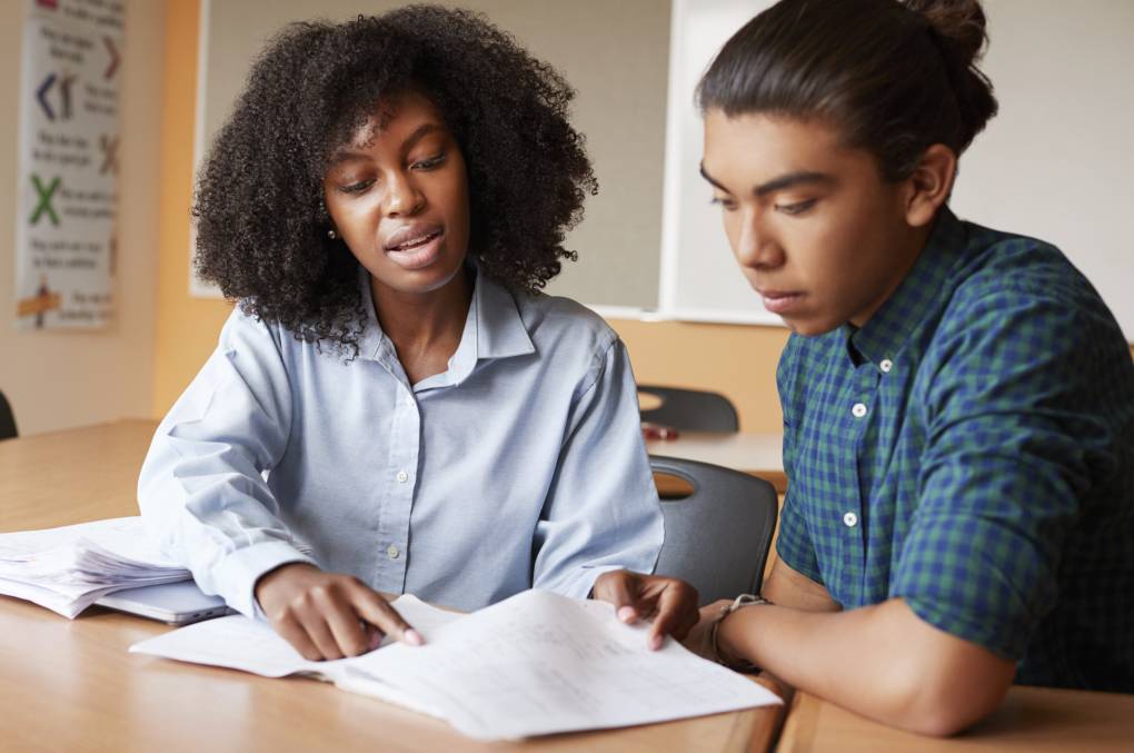 In a classroom, seated at students desks, a female tutor sits next to male high school student and points to a page in a workbook.
