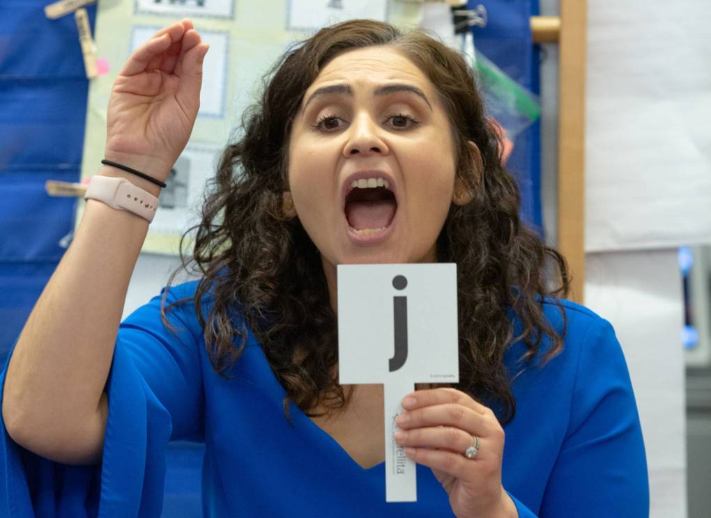A teacher in a blue shirt holds up the letter "j" her hand.