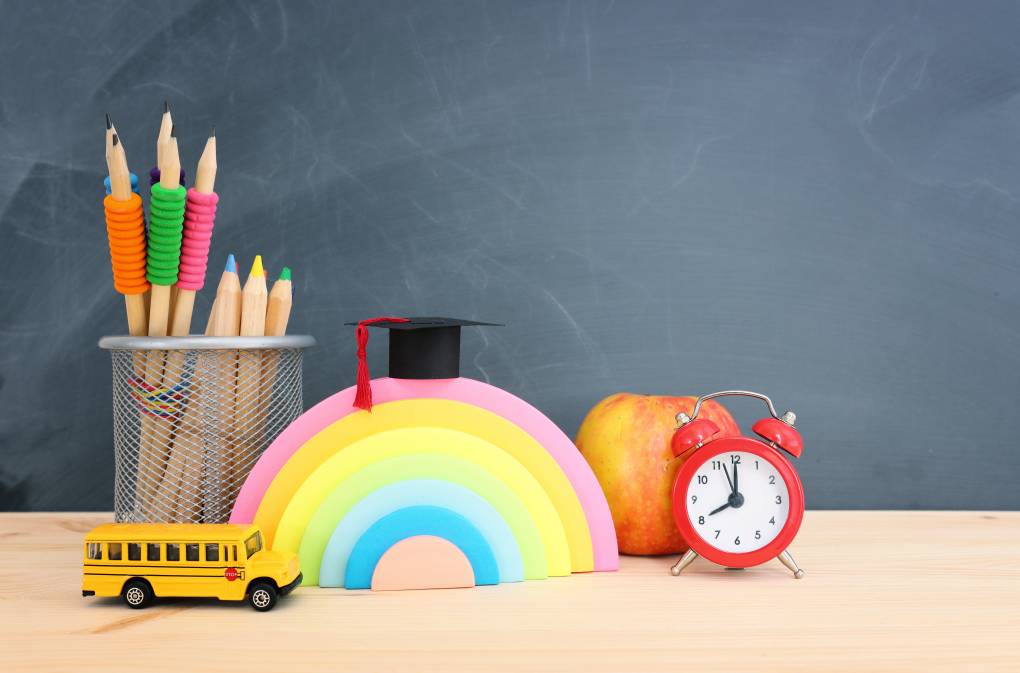 teacher desk surface with chalkboard in background. On the desk are a cup with colored pencils in it, a school bus figurine, a rainbow sculpture with a small graduation cap on it, an apple and a small, red alarm clock.