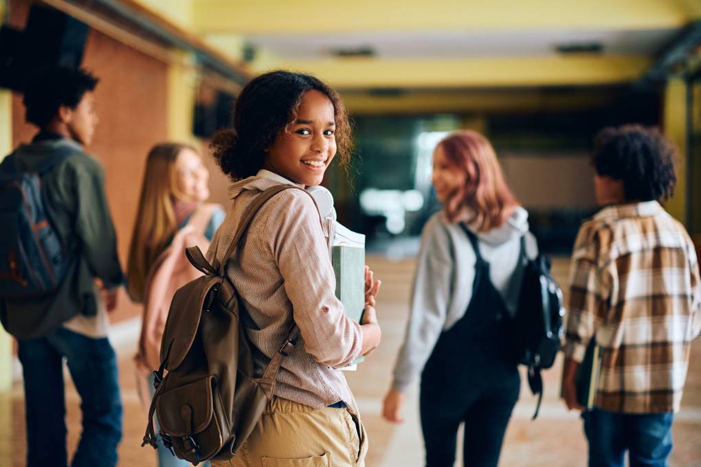 A Black young teenage girl walks through a school hallway wearing and backpack and looking back with a smile. Her friends walk a few feet ahead of her.