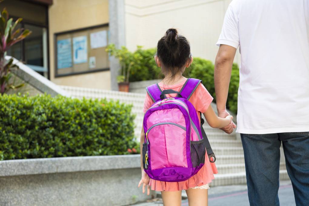 Father and young daughter both seen from behind as they approach the steps to a school. Girl wears a pink backpack and salmon colored dress. Her black hair is in a bun. Father wears white t-shirt and blue jeans.