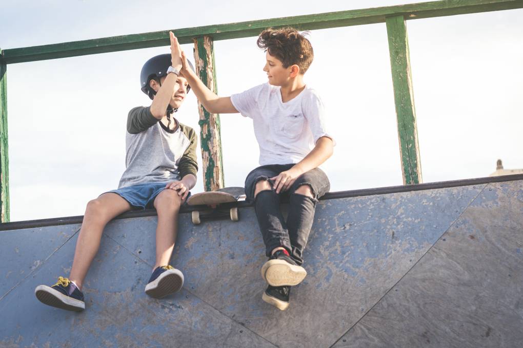 Two young boys hit high five. One boy wears a helmet. A skateboard rests between them ont he edge of a ledge. They are at the top of a structure in a skate park.