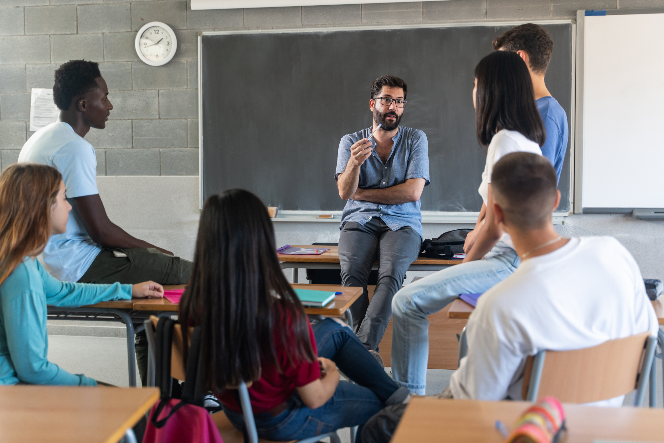 Teacher talking with teenager students sitting at and on desks.