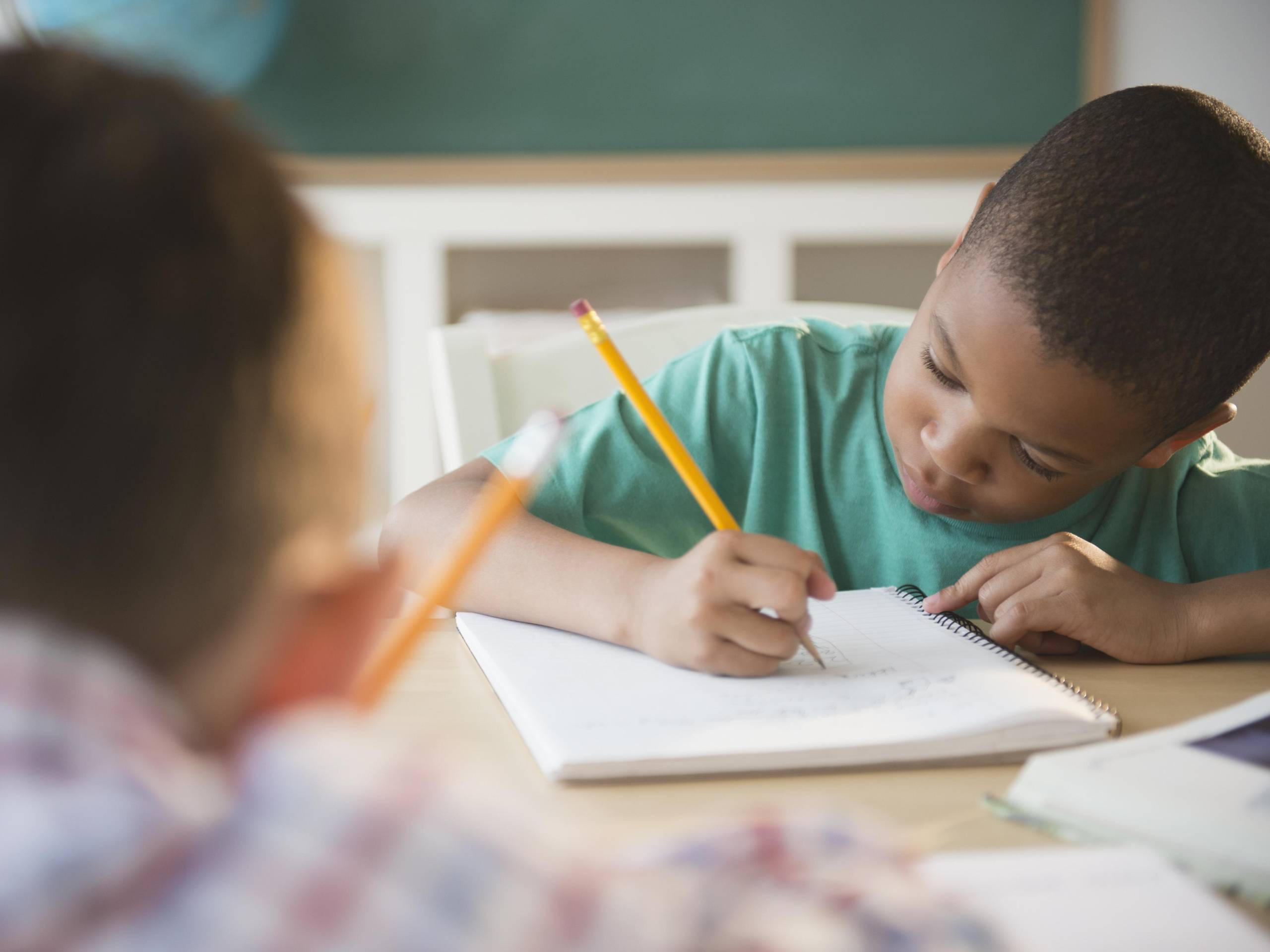 Two students writing at a desk with yellow pencils. The bottoom of a chalkboard is in the background.