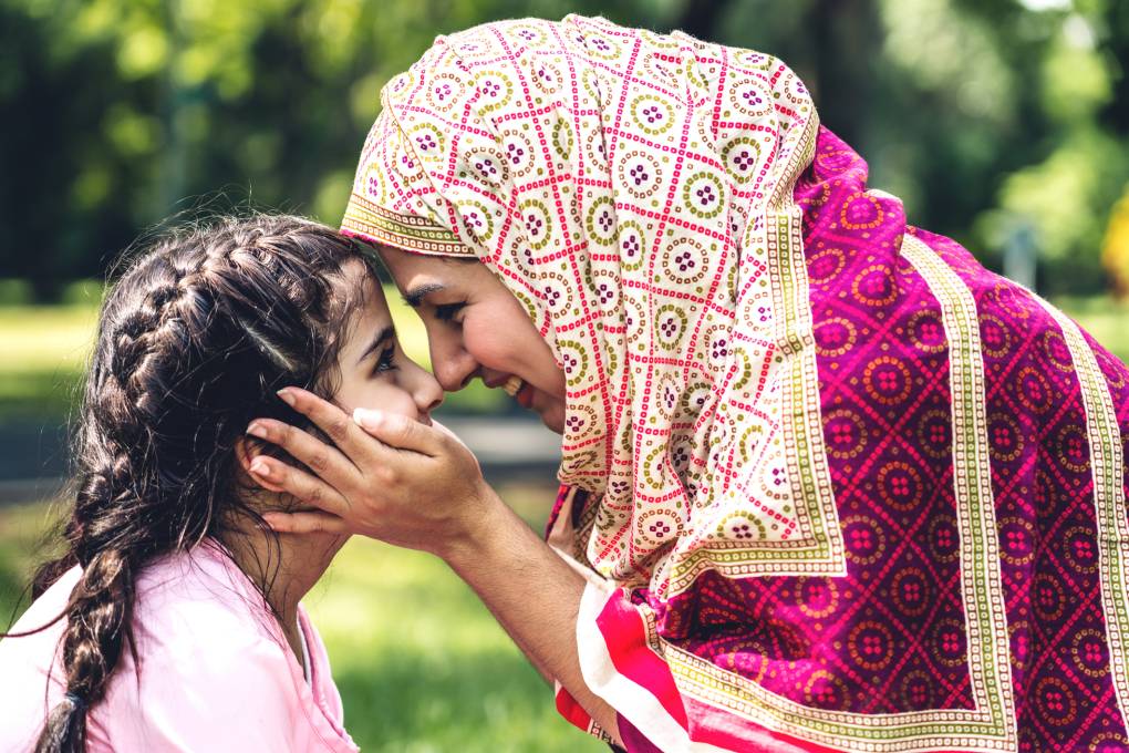 A child with two French braids and an adult woman wearing a patterned hijab face each other with smiles. The woman holds the child's face in her hands.