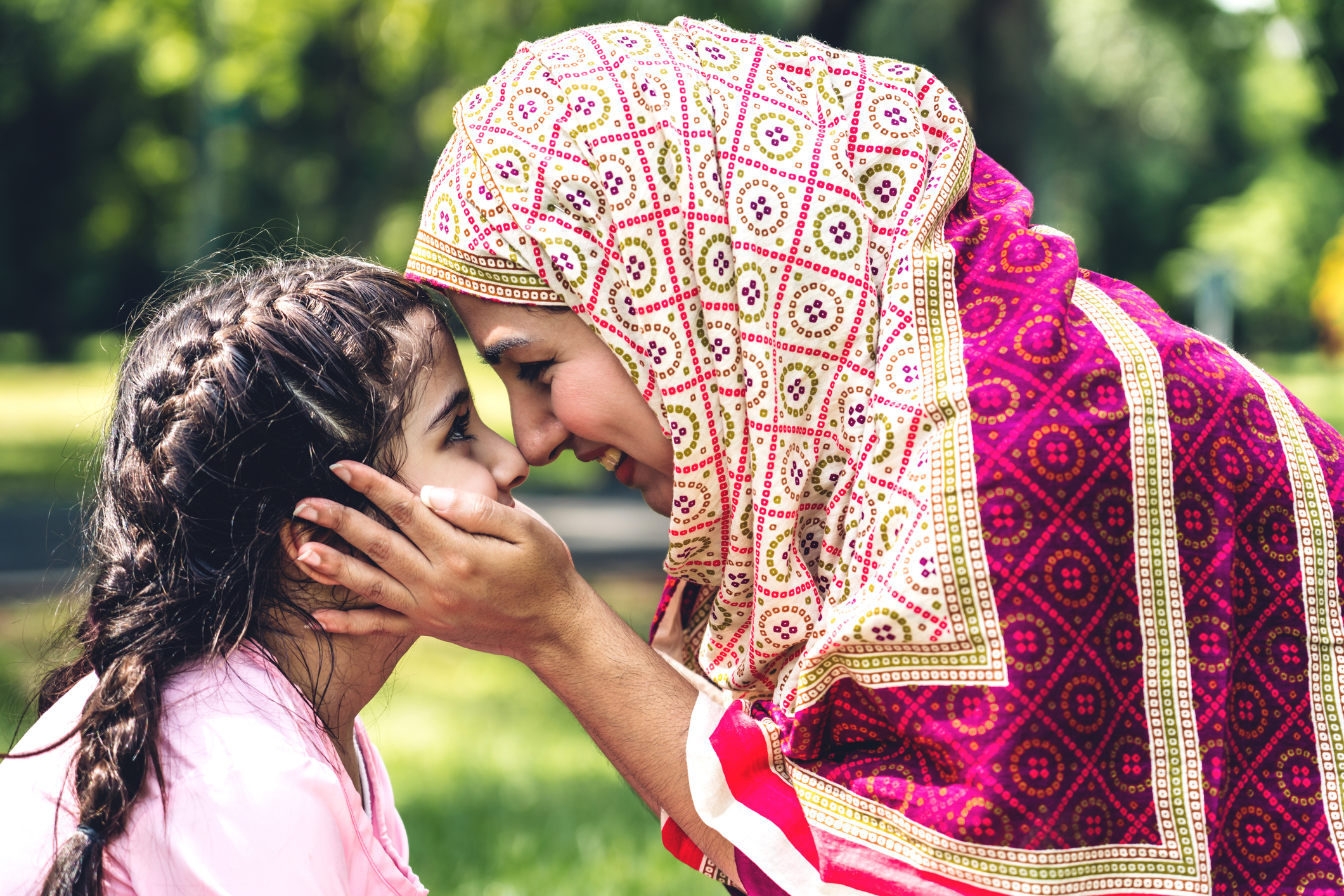 A child with two French braids and an adult woman wearing a patterned hijab face each other with smiles. The woman holds the child's face in her hands.
