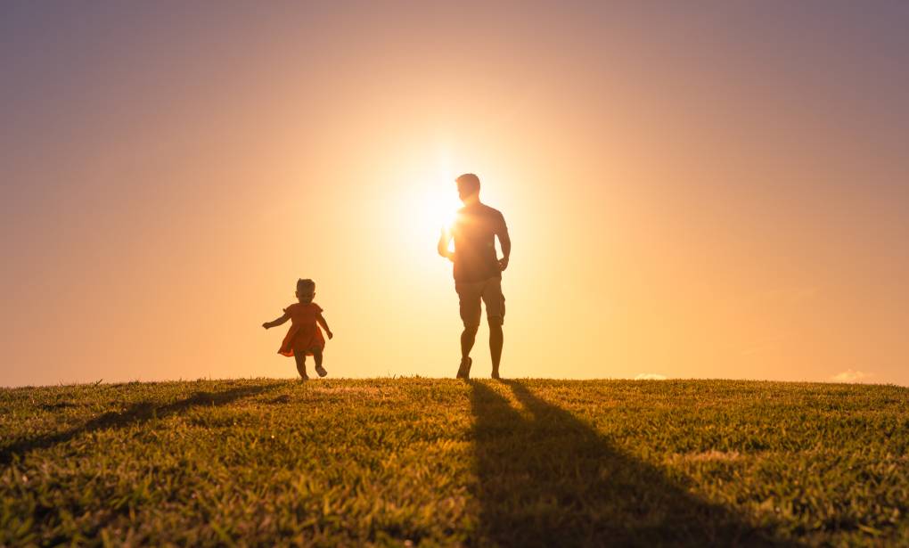 A father and young daughter stand on the horizon with sun setting behind them.