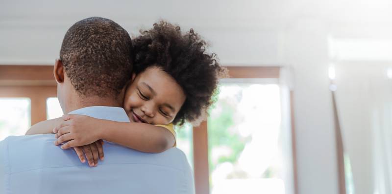 A Black father and child hug in a room with light shining through glass doors