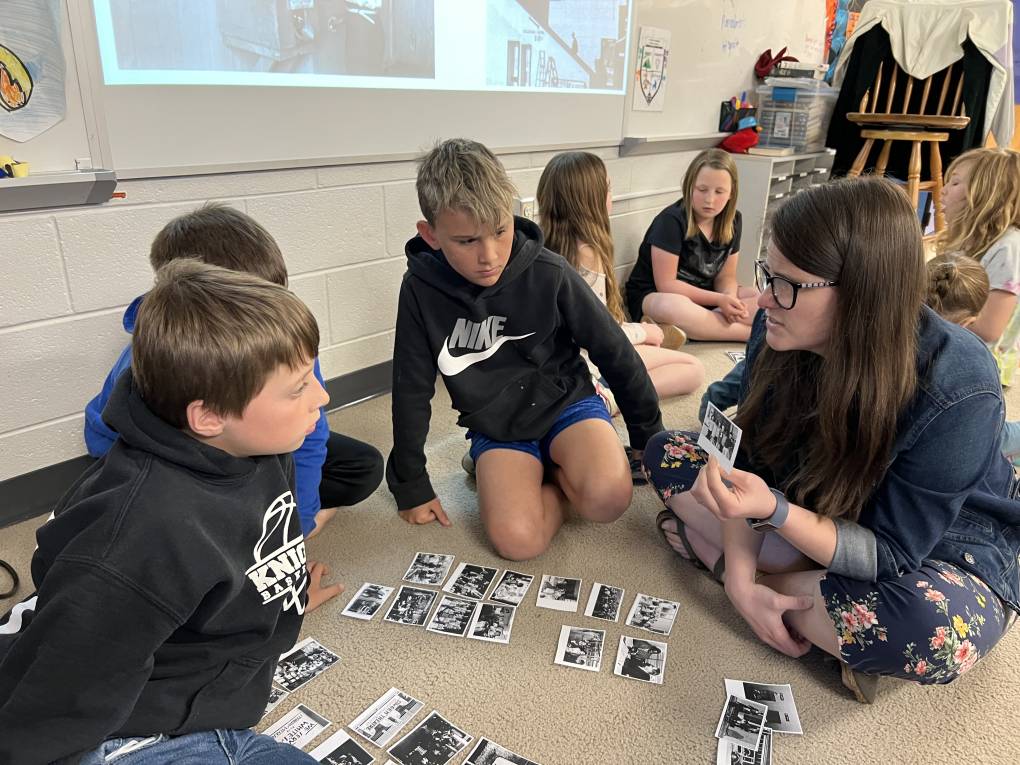 A teacher sits cross legged on a carpet in a circle with three young students. She holds and points to a printout of a black and white photo. Similar printouts are on the rug in the center of the circle. Another circle of students sit in the background.