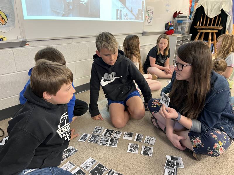 A teacher sits cross legged on a carpet in a circle with three young students. She holds and points to a printout of a black and white photo. Similar printouts are on the rug in the center of the circle. Another circle of students sit in the background.