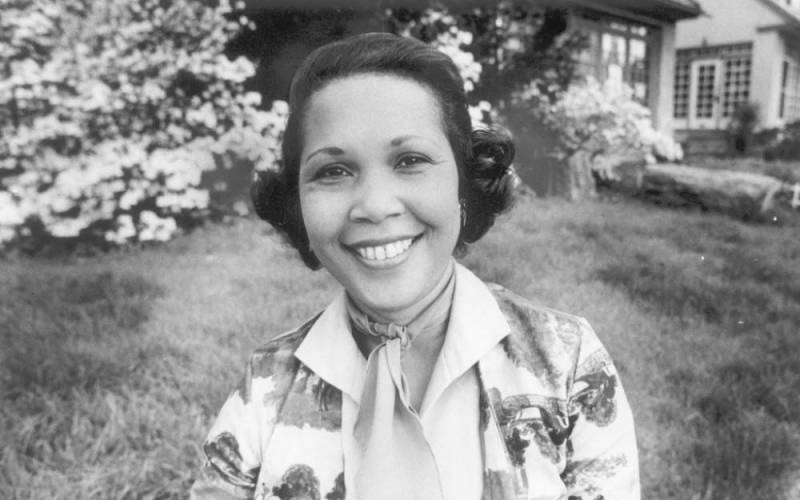 Black and white photo of a smiling woman shown from shoulders up. Grass, bushes and a house are in the background.