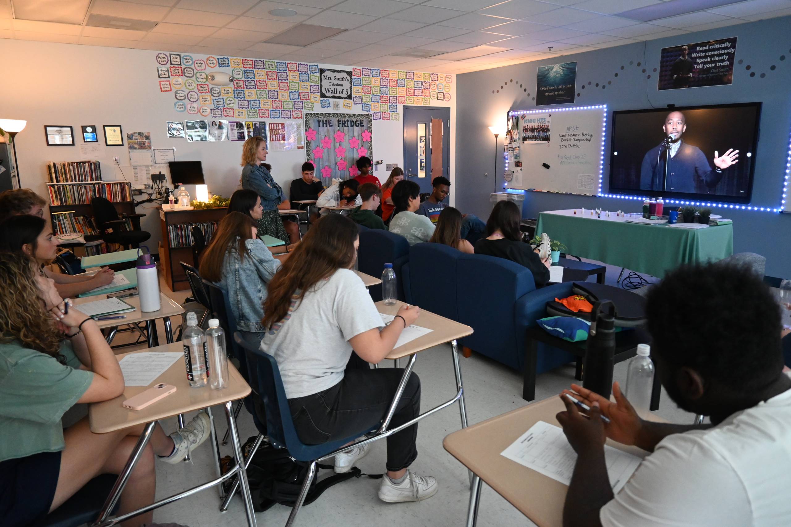 Students in desks and on a couch in a classroom take notes. A person appears on smartboard screen at the front of the room.